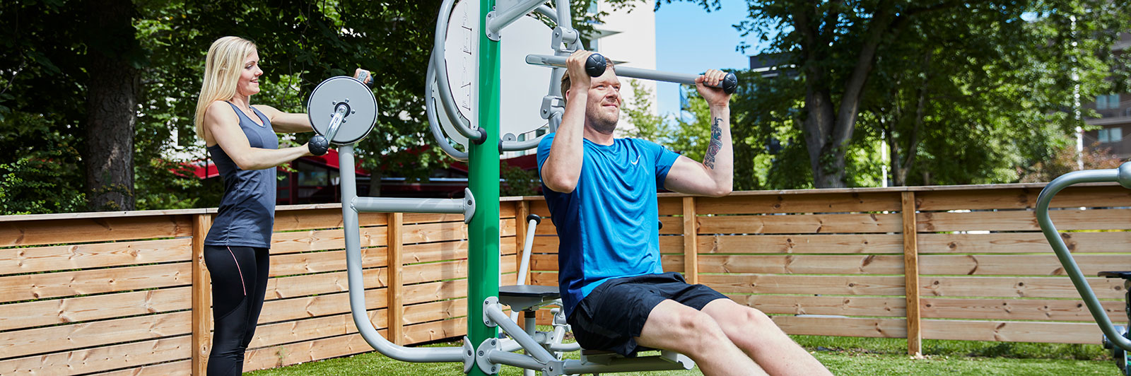 a man and woman exercise outdoors on various fitness machines.